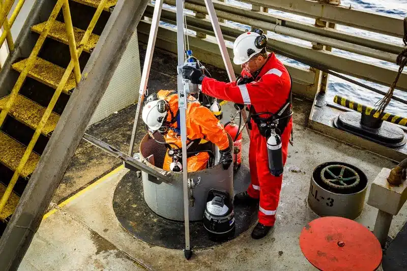 A confined space emergency rescue team in red and orange jumpsuits lowers one worker into a confined space entry point